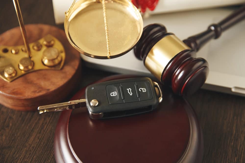 Image is of a gavel, balance scales, and a car key arranged together, concept of legal responsibility in a car accident case.