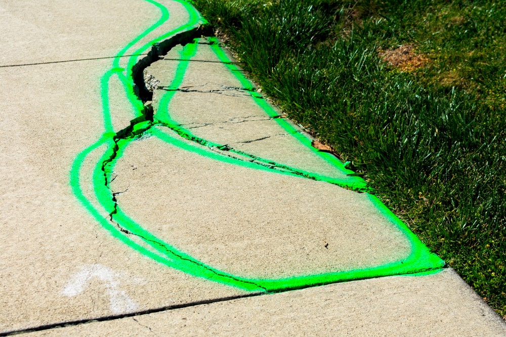 Image is of a severely cracked public sidewalk marked with bright paint, showing unsafe walking conditions and the risk of slip and fall injuries caused by sidewalk defects.