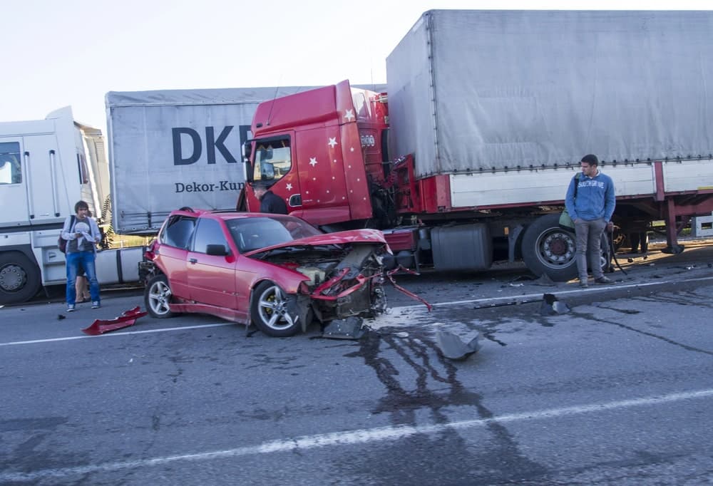 Image is of a damaged car wedged under a commercial truck after a collision, concept of fault in truck accidents and physical crash evidence.