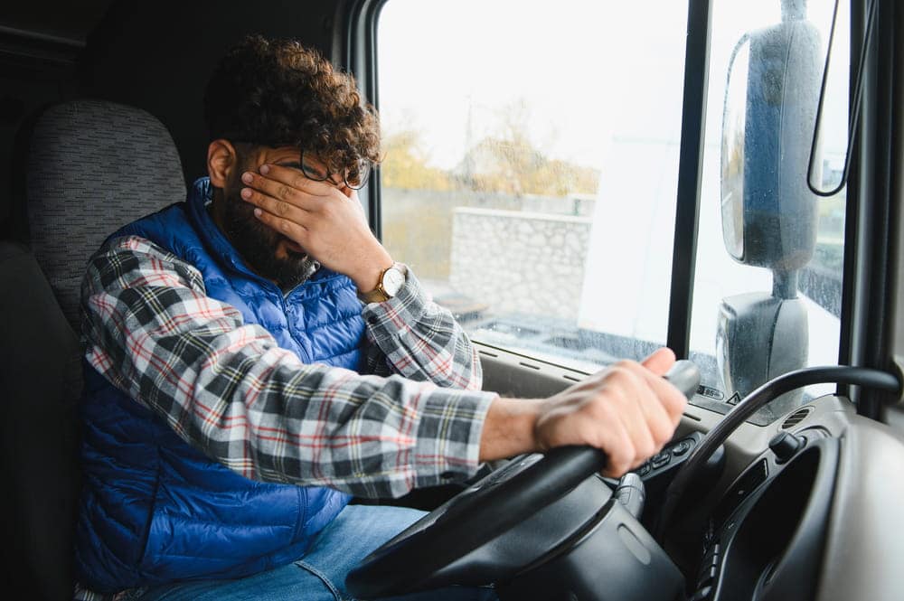 Image is of a fatigued truck driver sitting in a commercial truck cab covering his face, representing driver exhaustion that may contribute to a Nashville truck accident.