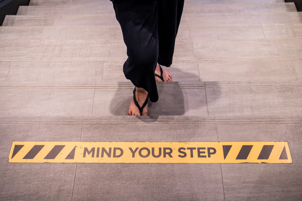 Image is of a person walking down stone stairs towards a yellow mind your step warning sign, illustrating how to prevent a slip and fall.