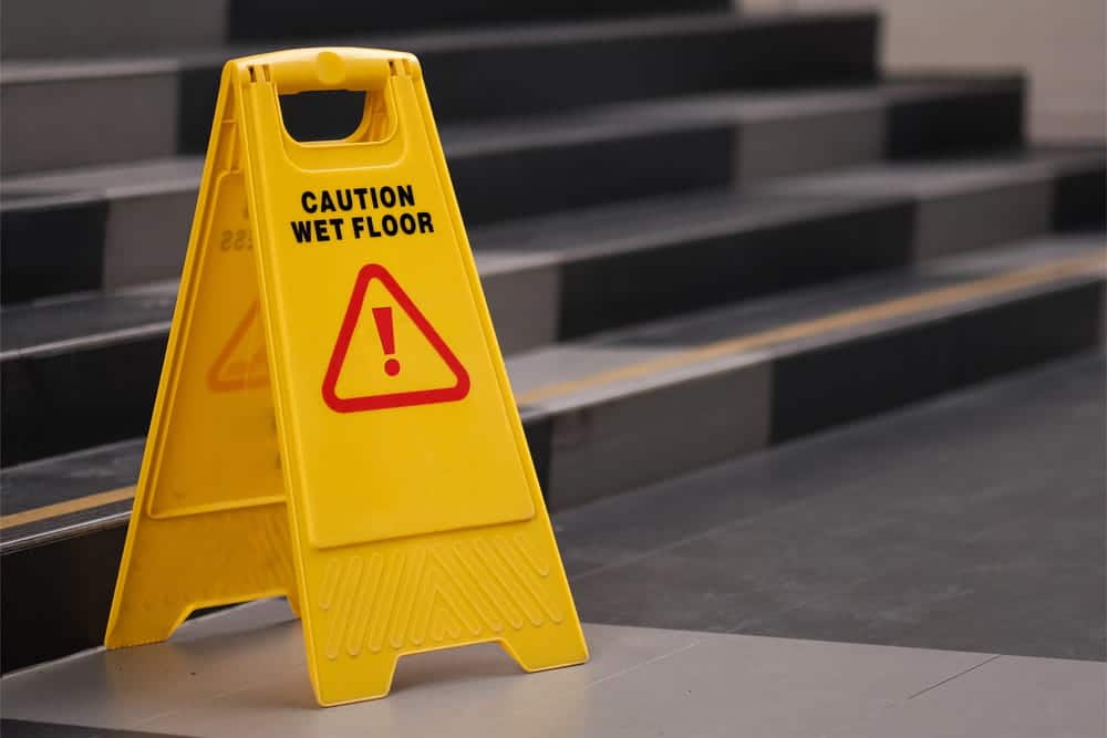 Image is of a bright yellow caution wet floor sign placed near a set of dark stairs, concept of a slip and fall hazard in a common area.