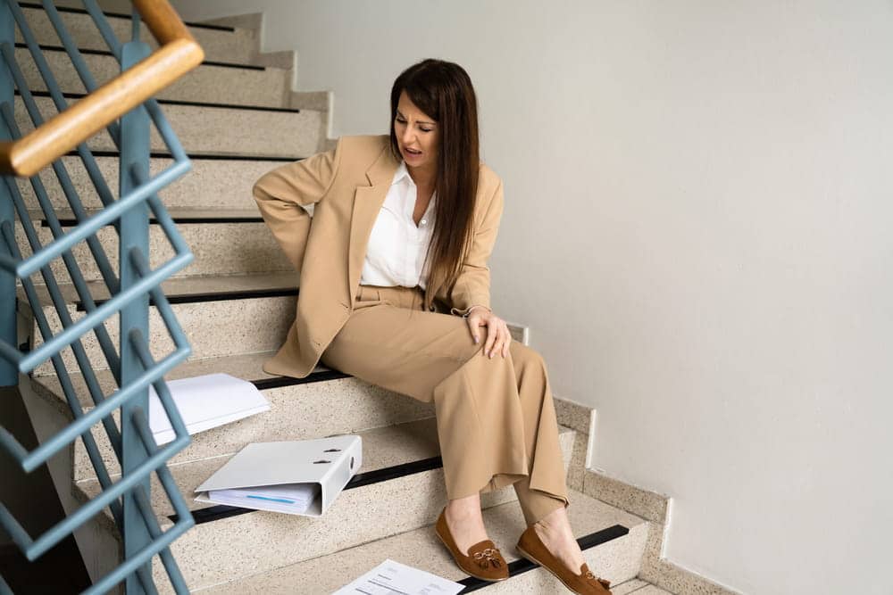 Image is of a woman sitting on stairs holding her back in pain, concept of slip and fall at work injury