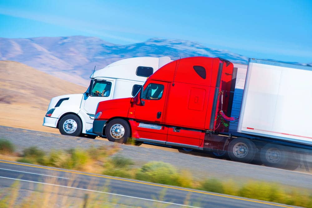 Image is of two semi trucks traveling side by side on a highway, concept of semi-truck blind spot risks while passing large trucks.