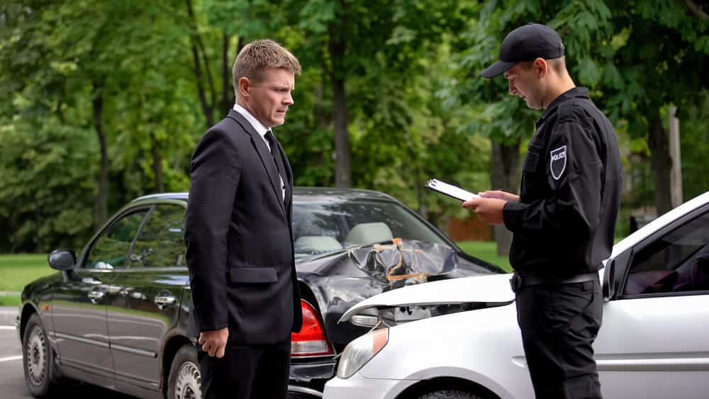 Image is of a police officer documenting vehicle damage while speaking with a driver after a car crash.