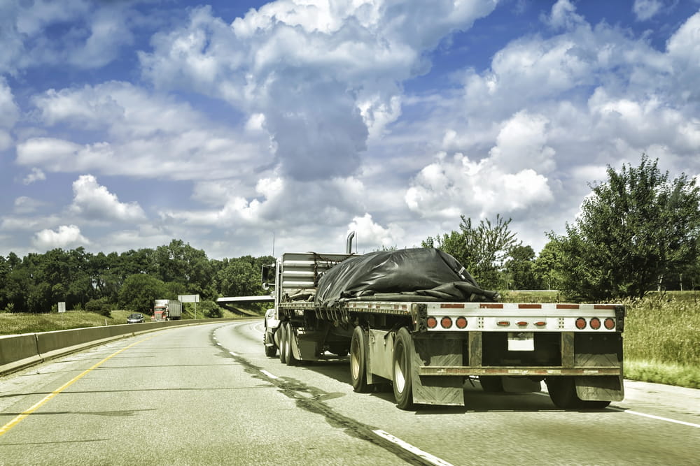 Image is of a flatbed truck transporting covered cargo on a highway, illustrating cargo securement issues that may affect a truck accident claim.